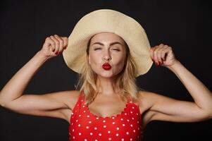 Close-up portrait of a beautiful sexy delightful stunning blonde European woman in red swimsuit with white polka dots and a straw summer hat sending a kiss, against black background with copy space photo