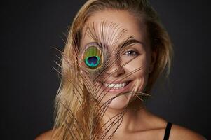 woman, with beautiful healthy glowing skin holding a peacock feather near her face. Black background copy space. photo