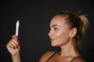 Side portrait of a beautiful blonde middle aged woman holding a tube with moisturizer, anti-aging beauty cosmetic product isolated over black background with copy space photo