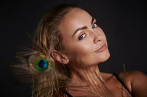 Headshot gorgeous smiling stunning middle aged blonde Caucasian woman with healthy fresh clean skin and wet blond hair holding a peacock feather, isolated over black background with copy ad space photo