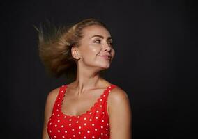 Smiling attractive stunning blonde woman with natural makeup and sun tanned skin wearing red swimsuit with polka dots and looking aside at a copy space on black background. Summer concept photo