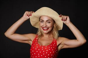 Attractive Caucasian blonde woman in a red swimsuit with white polka dots and a straw summer hat smiles a beautiful toothy smile, looking at the camera posing against black wall background. Copy space photo