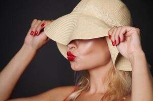 Close-up beautiful Caucasian woman covering her eyes with a straw summer hat and sending kiss, isolated over black background with copy space. Headshot portrait of a pretty lady with red lipstick photo
