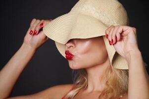 Close-up beautiful Caucasian woman covering her eyes with a straw summer hat and sending kiss, isolated over black background with copy space. Headshot portrait of a pretty lady with red lipstick photo