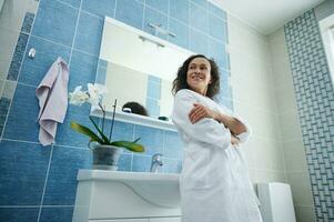 mujer en bata de baño posando con cruzado brazos en frente de el cámara propensión en el lavabo en su hogar baño foto
