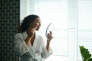 Beautiful woman wearing a bathrobe uses a jade Gua Sha massager on her cheek, looking into her mirror reflection while standing in a bathroom with a window in the background photo