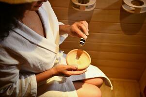 Focus on hands. Top view of a woman in bathrobe steamed in sauna and pouring some drops of essential oil in wooden mortar photo