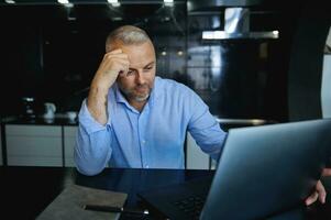 Pensive handsome mature Caucasian businessman, entrepreneur sitting in front of a laptop looking thoughtfully at screen monitor sitting at table against a stylish home kitchen background photo