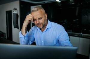 perplejo pensativo hermoso medio Envejecido europeo empresario, empresario sentado en frente de un ordenador portátil mirando pensativamente a pantalla monitor sentado a mesa en contra un elegante hogar cocina antecedentes foto