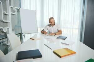 Handsome middle aged Caucasian man sitting at a table near a flip chart with copy ad space and writing, making notes on notepad photo