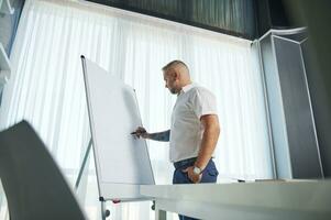 Side portrait of a middle aged Caucasian man in white casual shirt and tattooed arms writing business plan on flip chart photo