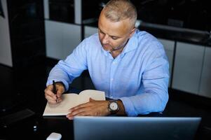 Top view of a middle-aged European man writing in a notebook, planning projects, and making notes in a diary, sitting in front on laptop at home office photo