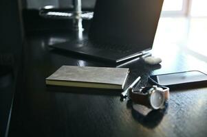 Close-up view of office accessories, laptop, earphones on the box, watch, mobile phone and gray notepad on wooden desk photo