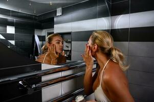 Charming young Caucasian blonde woman in swimsuit applying moisturizer, performing smoothing rejuvenating facial massage looking at her reflection in bathroom mirror photo