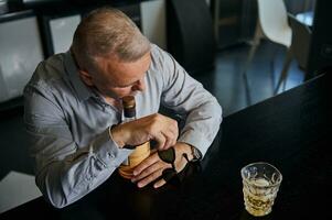 Handsome tired exhausted mature businessman resting after work , sitting at a table and drinking alcoholic drink at home kitchen bar photo