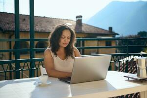 confidente hembra real inmuebles agente trabajando en ordenador portátil desde hogar balcón, en contra un hermosa paisaje de italiano Alpes foto