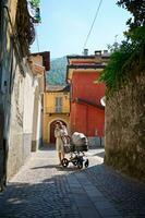 Full length portrait of a pretty woman in casual dress, standing with her baby in stroller in medieval cobblestone alley photo