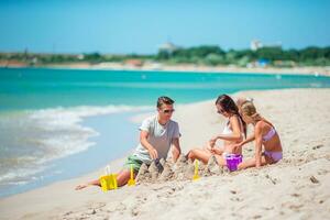 padres y pequeño hija disfrutando hora en el playa. familia haciendo arena castillo juntos en el costa foto