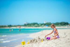 pequeño niña a tropical blanco playa haciendo arena castillo y jugando con playa juguetes foto