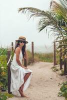 Happy woman on her way on the beach. Smiling girl in white beach dress and straw hat on summer tropical vacation photo