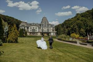 The bride and groom run through a picturesque field on the wedding day in the background of a wonderful forest and expensive hotel photo