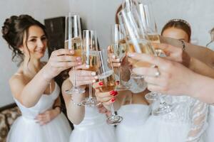 novia con alegre novias a el Boda bebida champán desde lentes. novia y novias abrazo en el habitación. Mañana novia y novias. blanco vestido. foto