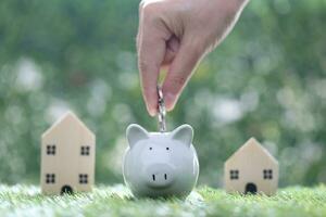 Woman hand putting a coin into piggy bank with model house on nature green background,Save money for prepare in future and family finance concept photo