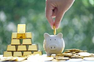 Woman hand putting a coin into piggy bank and gold bar on natural green background,Business investment and Saving money for prepare in future concept photo