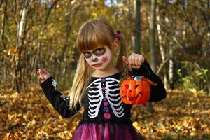 pequeño niña en Víspera de Todos los Santos esqueleto negro vestido, con azúcar cráneo maquillaje de Calavera catrina mascarada disfraz para día de muerto, dia Delaware los muertos. atención en naranja calabaza el plastico Cubeta con dulces. foto