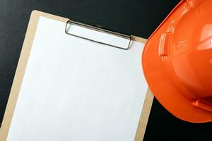 Blank clipboard paper and hard hat or safety helmet isolated on black background. photo