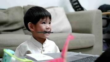 ásia Garoto detém lápis dentro boca e mostra entediado expressão enquanto estudando conectados em computador portátil video