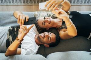 Portrait of laughing couple lying on the the couch taking selfies with smartphones photo