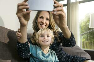 sonriente madre con hijo tomando un selfie en sofá a hogar foto