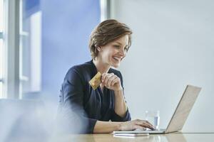 Smiling businesswoman holding credit card and using laptop at desk in office photo