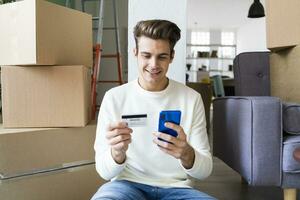 sonriente joven hombre pago mediante móvil teléfono por crédito tarjeta mientras Moviente en nuevo casa foto