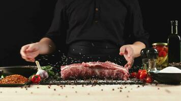 Koch bewirbt sich gemasert Salz- auf roh Stück von Steak. Kocher vorbereiten Fleisch auf Fachmann Küche Tabelle mit verschiedene Gemüse 4k Aufnahmen video