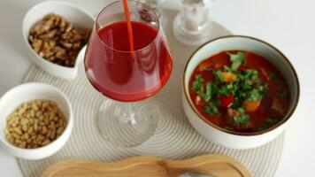 Tomato juice pouring into a glass. Soup with beef, potatoes, carrot, tomatoes, paprika, onion and bell peppers. White bowl on white table. video