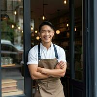 retrato de contento asiático joven hombre en pie a puerta de su almacenar. alegre maduro camarera esperando para clientela a café tienda. pequeño negocio dueño. generativo ai foto