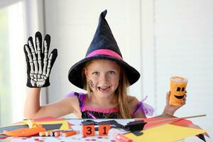 A little girl in a witch costume looks at the camera and laughs, holding a glass pumpkin and a skeleton glove in her hands. photo