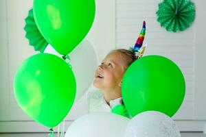 pequeño niña con un unicornio aro en su cabeza en contra el antecedentes de verde globos cumpleaños para niños. foto