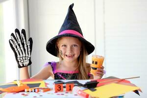 A little girl in a witch costume looks at the camera and laughs, holding a glass pumpkin and a skeleton glove in her hands. photo