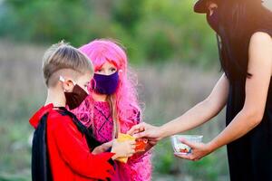 niños truco o tratar en Víspera de Todos los Santos disfraz y médico mascarilla. un chico y un niña en trajes durante el coronavirus pandemia recibir caramelo desde un mujer . foto