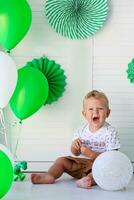 A little boy sits on the floor against the background of green balloons and decorations. Birthday for children. Celebrating St. Patrick's Day. photo