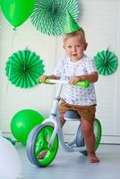 A little boy with a cap on his head sits behind a balance bike against the background of green balloons. Birthday for children. Celebrating St. Patrick's Day. Gift bike for kid photo