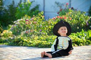 Little boy  sitting on the grass wearing a skeleton costume and a black hat. Halloween holiday. photo