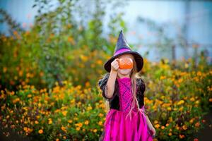un pequeño niña vestido como un bruja soportes en el calle y se esconde su cara con un calabaza para dulces foto