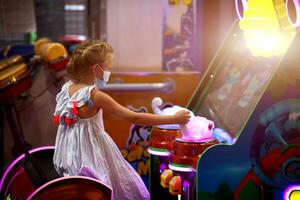 A little girl in a medical mask plays a game machine in the children's room. photo