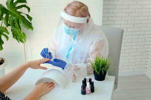 Manicurist in are wearing PPE paints nails in a beauty salon. Professional procedure. The existence of small businesses in isolation from COVID-19. photo
