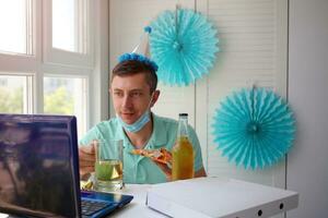 A man in a medical mask sits in front of a laptop and waves hello. Celebrates her birthday via the internet in quarantine time, online birthday. photo