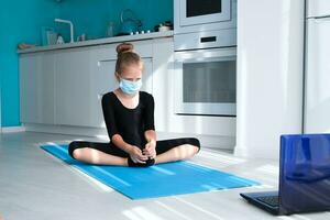 Fit little girl in medical mask doing yoga and watching online lessons on laptop, exercising in the kitchen. photo